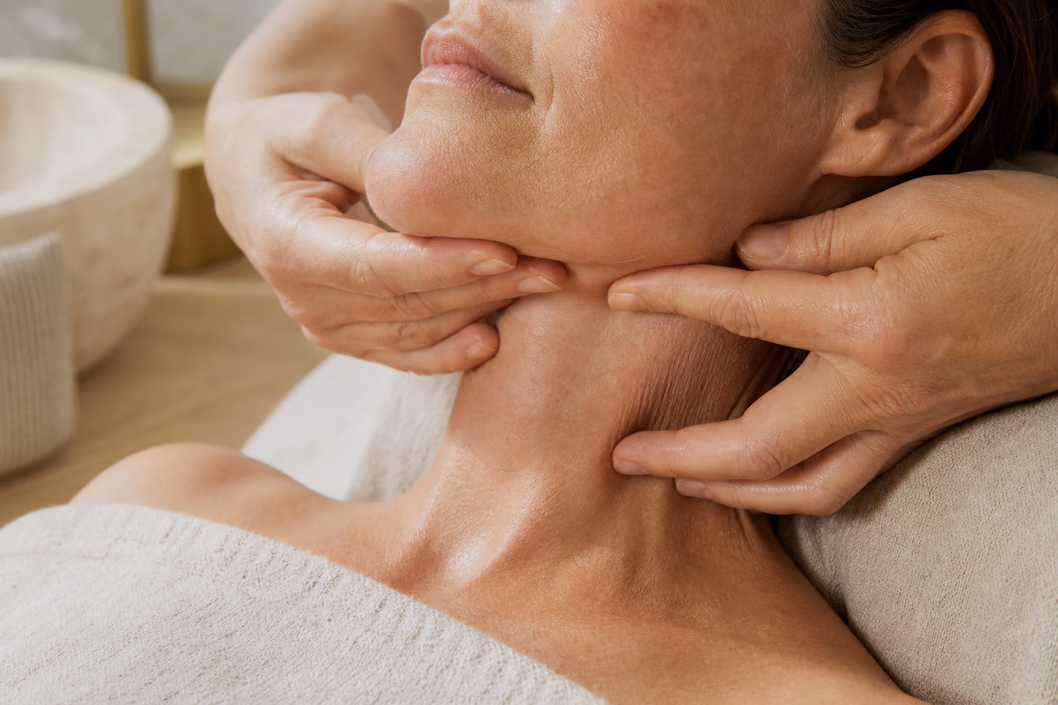 An esthetician's hands lifting the skin along a mature client's jawline during a collagen facial.
